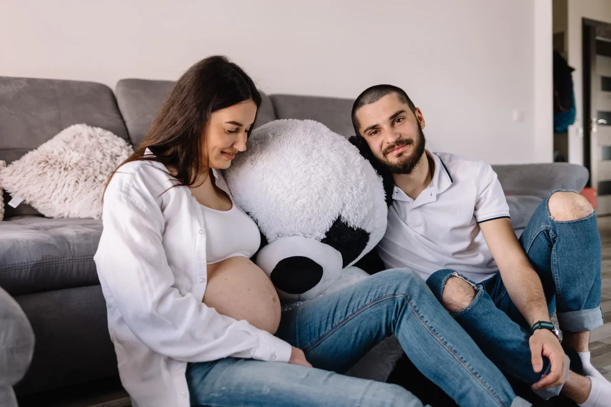 Schwangere Frau sitzt entspannt auf dem Sofa neben ihrem Partner und hält ihren Babybauch, beide wirken ruhig und zuversichtlich